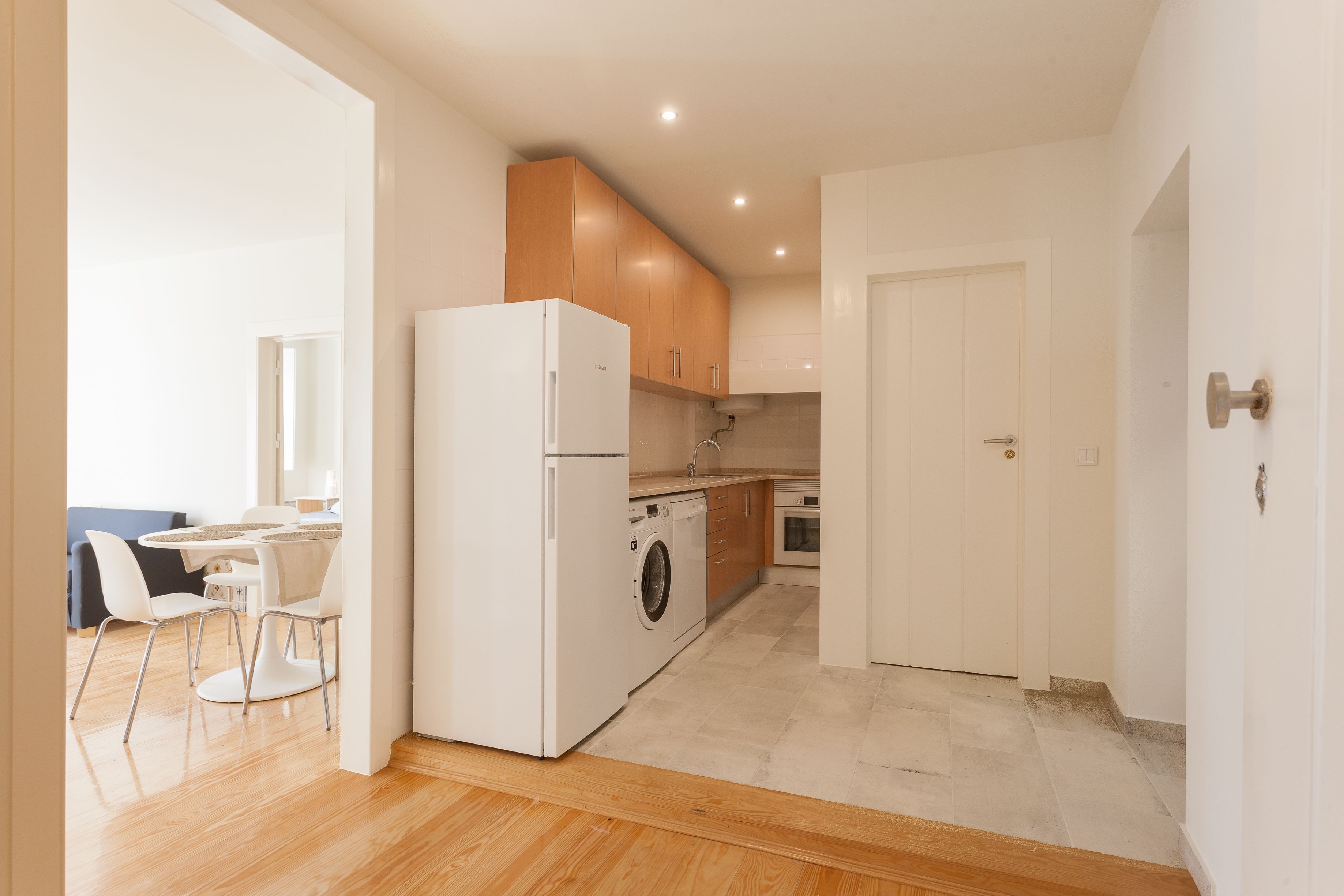 A modern kitchen with wooden cabinets, a built-in washing machine, and a dining area with white chairs and a table.