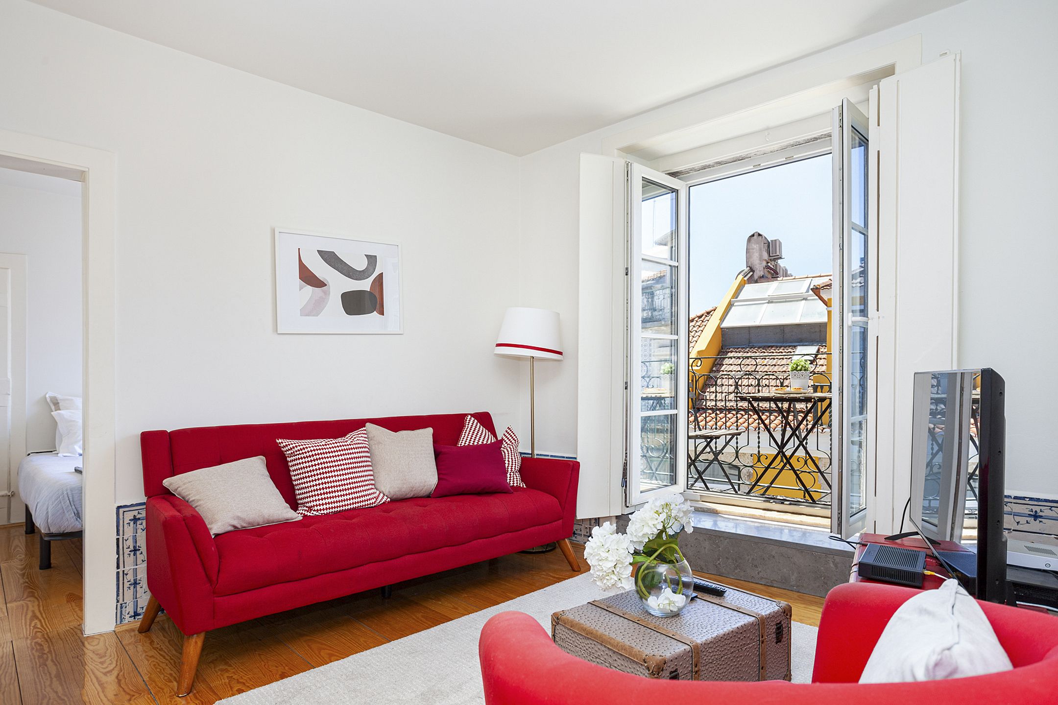 A red sofa with white and red cushions, a white lamp, a television, and a view of a balcony with a yellow railing.