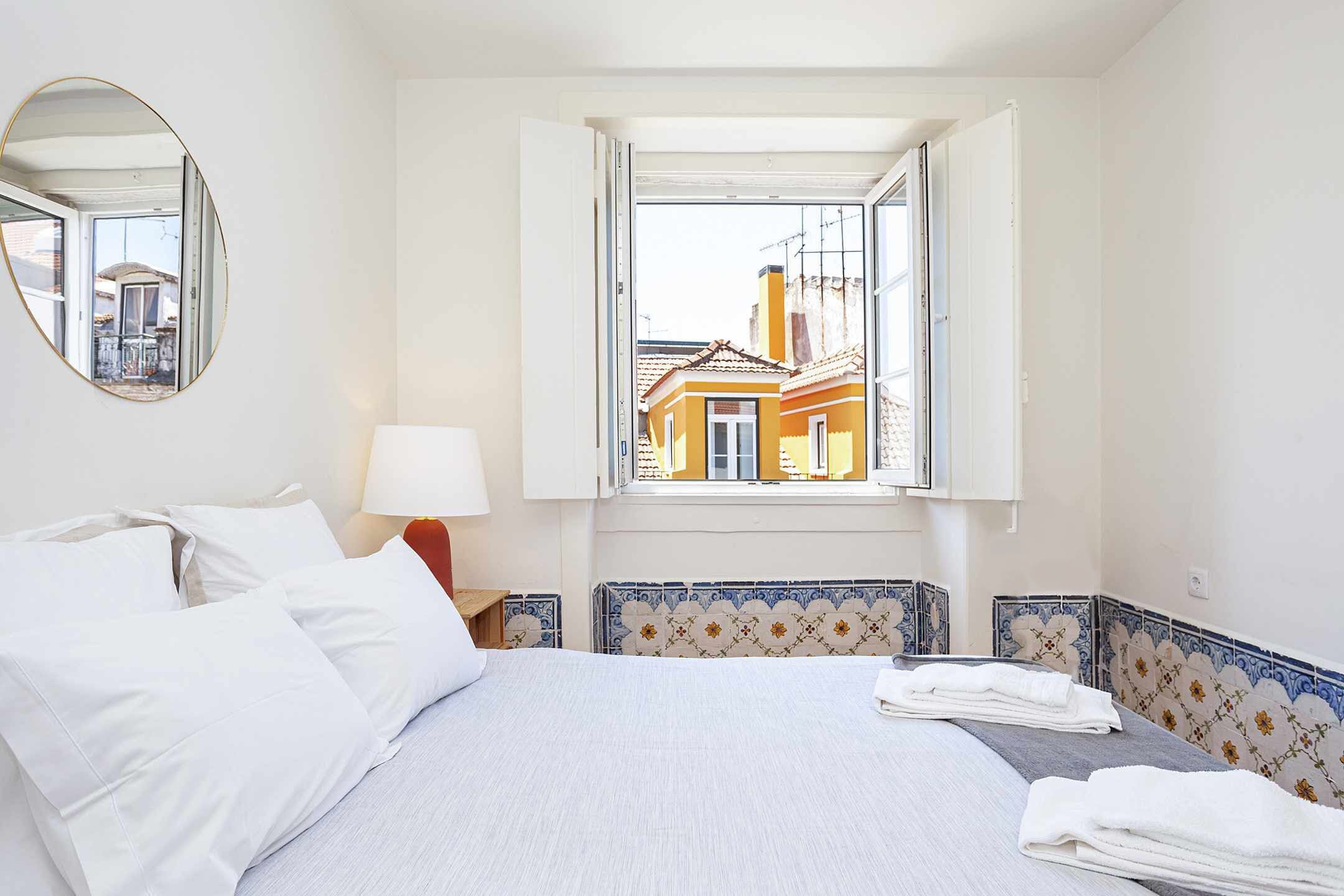 Una habitación de hotel con una cama, un ventanal con vistas a edificios, un espejo redondo y una lámpara.