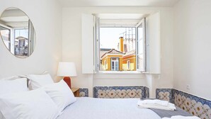Hotellihuone, jossa on sänky, ikkuna, josta on näkymä rakennuksiin, pyöreä peili ja lamppu.
