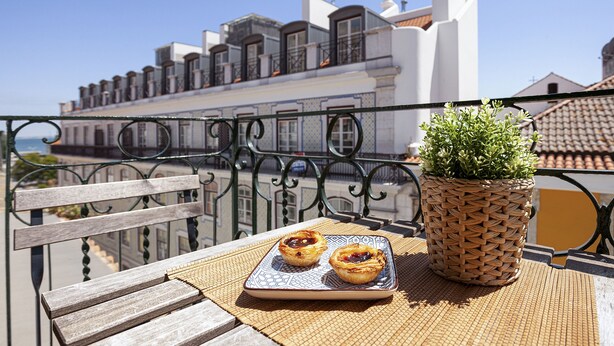 A balcony with a wicker pot of green plants, two pastries on a tray, and a view of a building with white and red roofs. - Gonzalo's Guest Apartments - Downtown Historic Flats III (Lisbon)