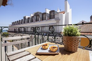 A balcony with a wicker pot of green plants, two pastries on a tray, and a view of a building with white and red roofs. - Gonzalo's Guest Apartments - Downtown Historic Flats III (Lisbon)