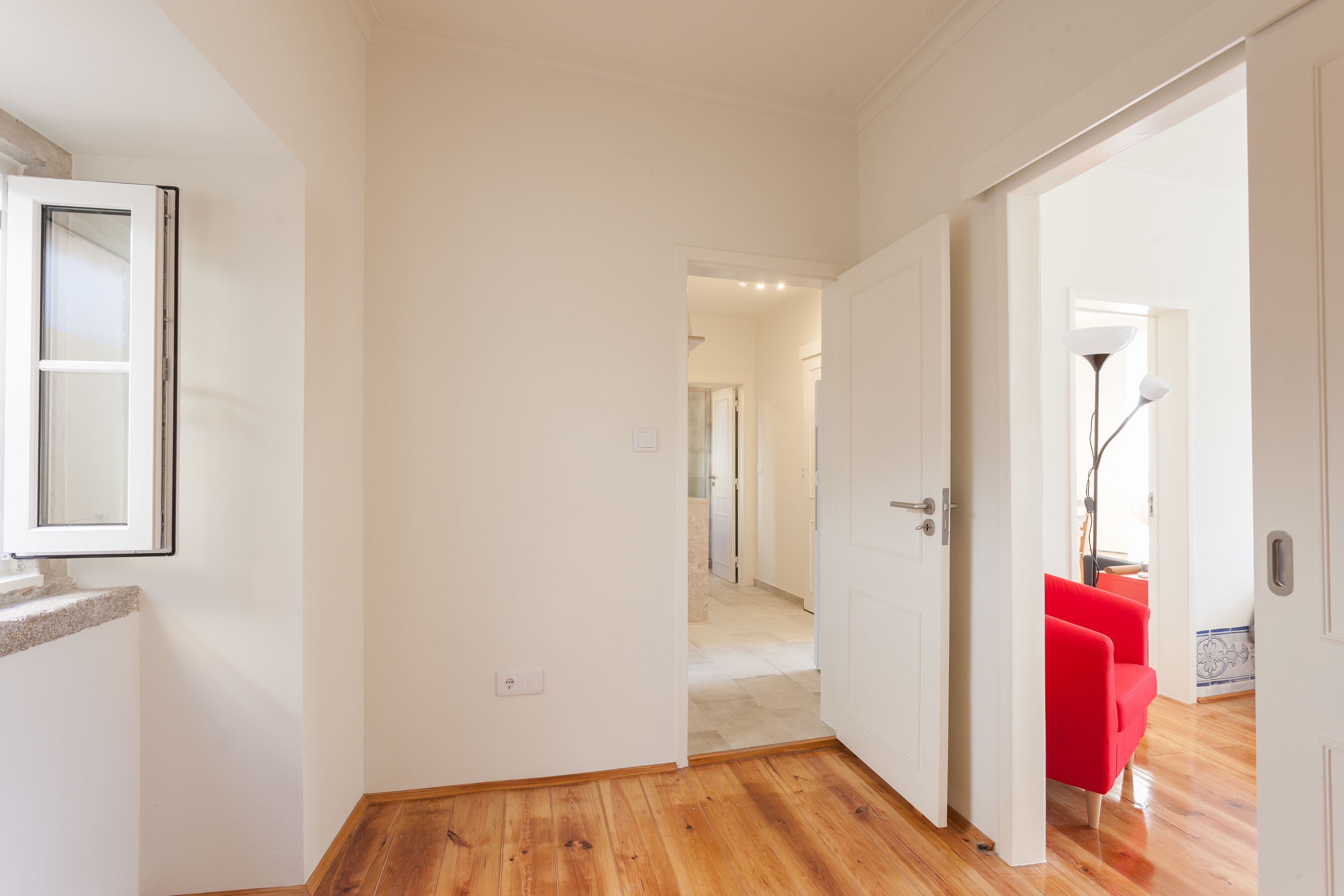 A hallway with wooden flooring, white walls, and a red chair in the distance.