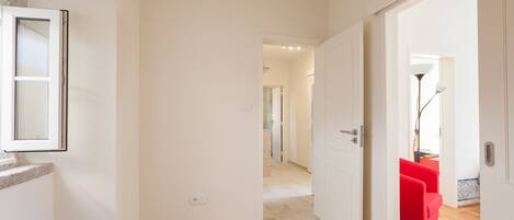 A hallway with wooden flooring, white walls, and a red chair in the distance.