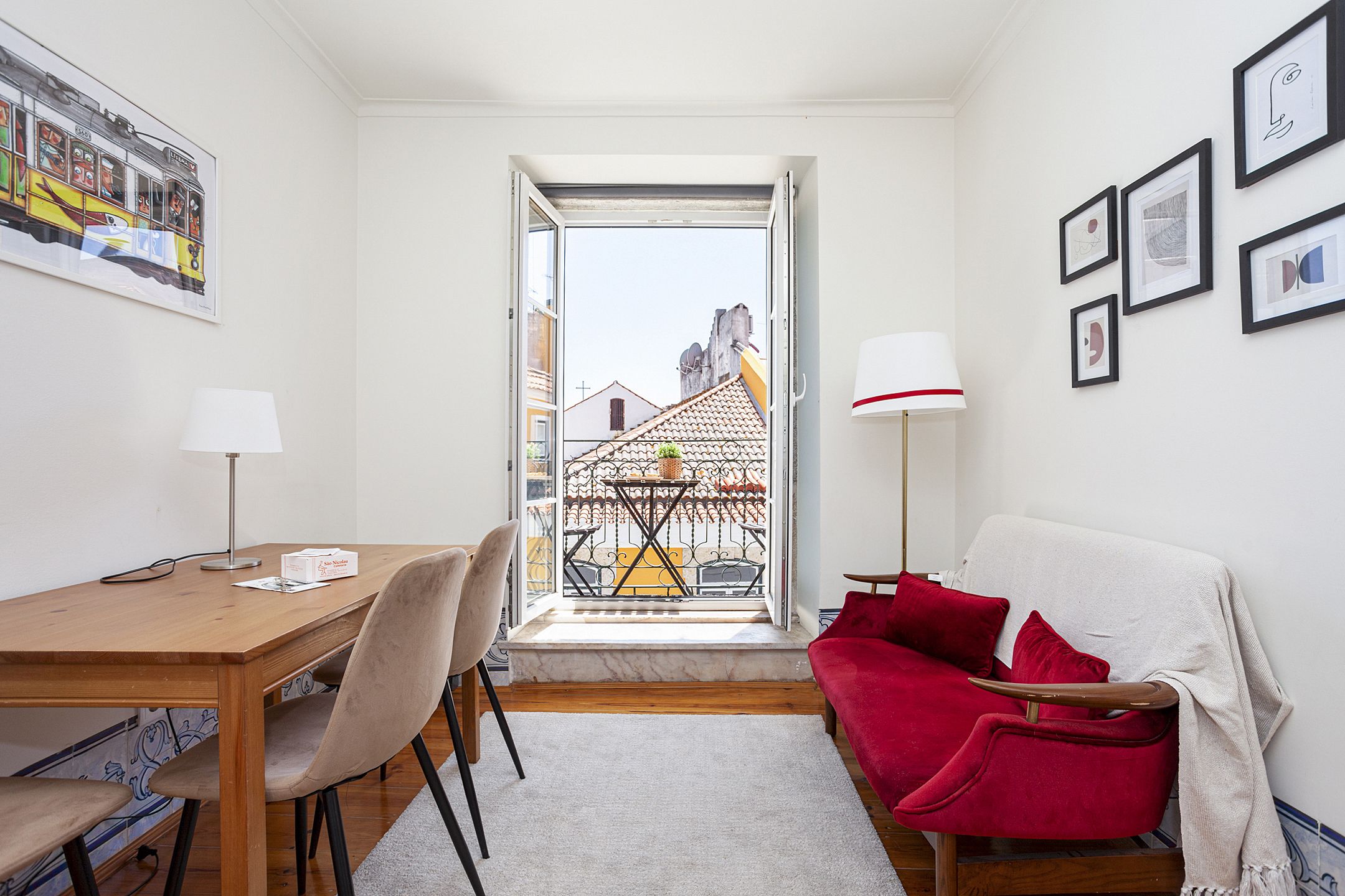 A compact room with a wooden table, two chairs, a red sofa, a white lamp, and a balcony with a view of buildings.