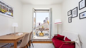 A compact room with a wooden table, two chairs, a red sofa, a white lamp, and a balcony with a view of buildings. - Gonzalo's Guest Apartments - Downtown Historic Flats III (Lisbon)