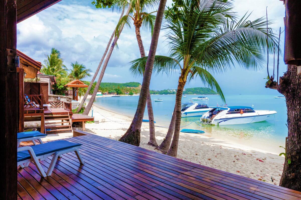 Eine Holzterrasse mit Liegestühlen, Blick auf einen Strand mit Booten, Palmen und blauem Himmel.