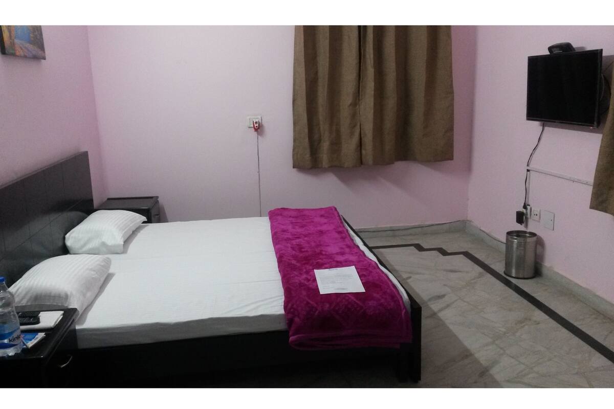 Een eenpersoonsbed met witte en roze beddengoed, een bruin gordijn, een aan de muur bevestigde televisie en een prullenbak.