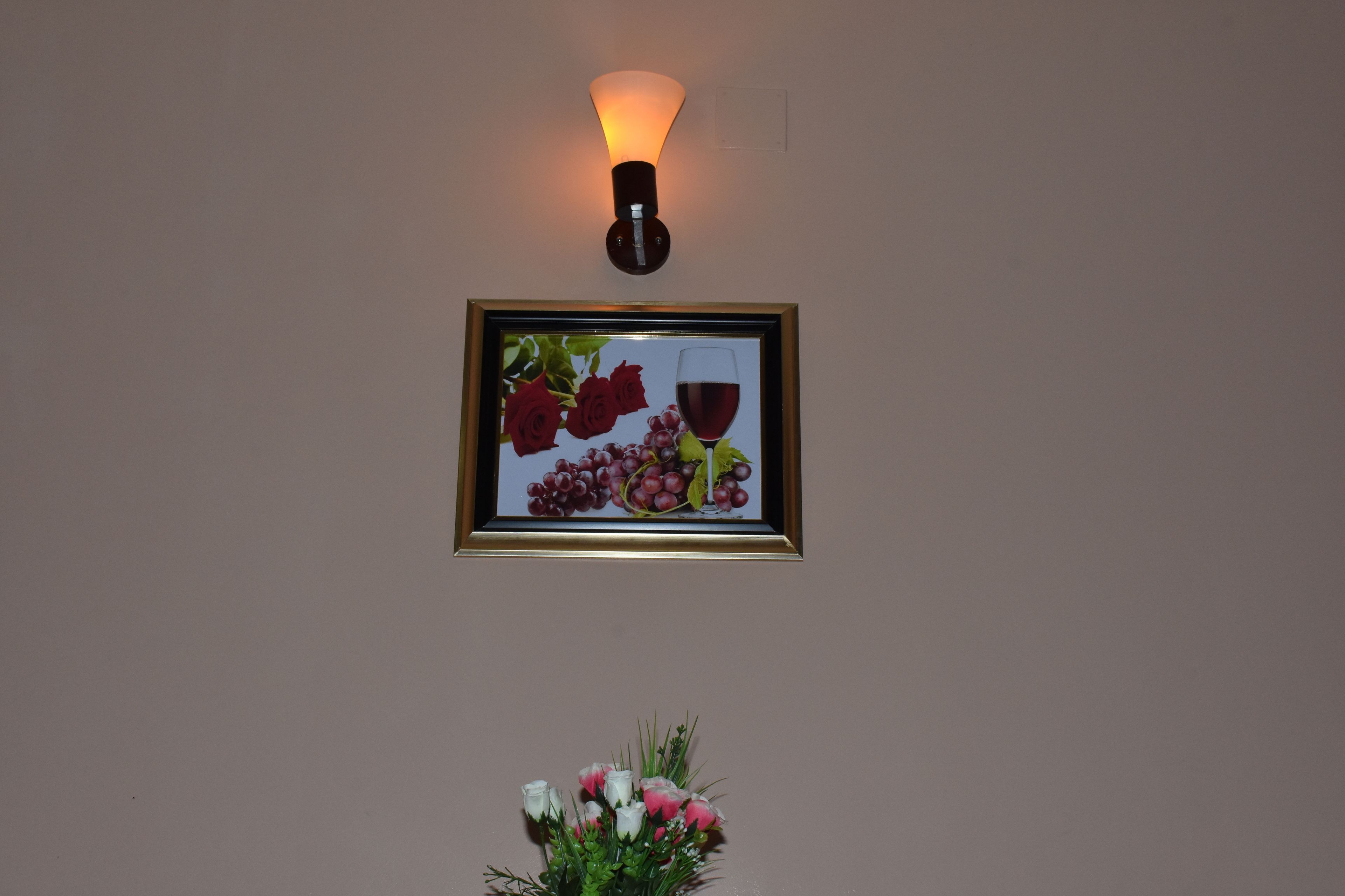 Ein gerahmtes Kunstwerk mit Trauben und Rosen an einer Wand, eine Wandleuchte und darunter ein Blumenvasen.