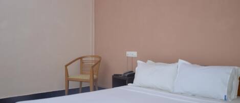 A single bed with white linens, a wooden chair, a bedside table with a lamp, and a wall socket.