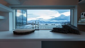 Ett rum med ett stort fönster som ger utsikt över ett snötäckt landskap med berg. Där finns ett träbord med ljus och en filt på.