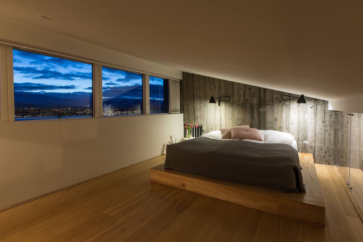 Un dormitori modern amb un llit gran, vista a un paisatge urbà a la posta de sol, i una paret d'accent de fusta.