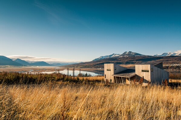 A modern building with a wooden exterior, situated in a grassy field with mountains in the background. - Villa Lola (Akureyri)