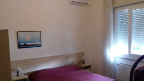 Un dormitorio con cama, mesita de noche, aire acondicionado instalado en la pared y ventana con persianas.