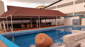 A swimming pool with a tiled edge, a covered seating area with tables, and a building with a grid-like facade in the background. - Golden Hôtel (Abidjan)