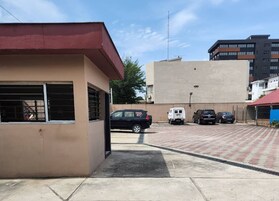 Un estacionamiento con varios autos, un edificio de techo rojo y una construcción color beige con ventanas protegidas por rejas.