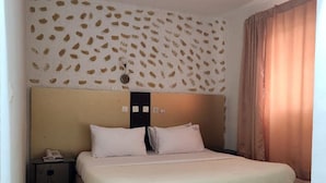 A single bed with a headboard, a bedside table with a phone, and a wall with a leaf pattern. - Golden Hôtel (Abidjan)