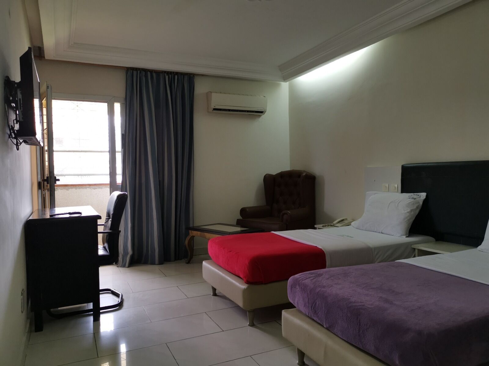 A hotel room with two beds, a desk, a chair, an air conditioning unit, and a window with curtains.