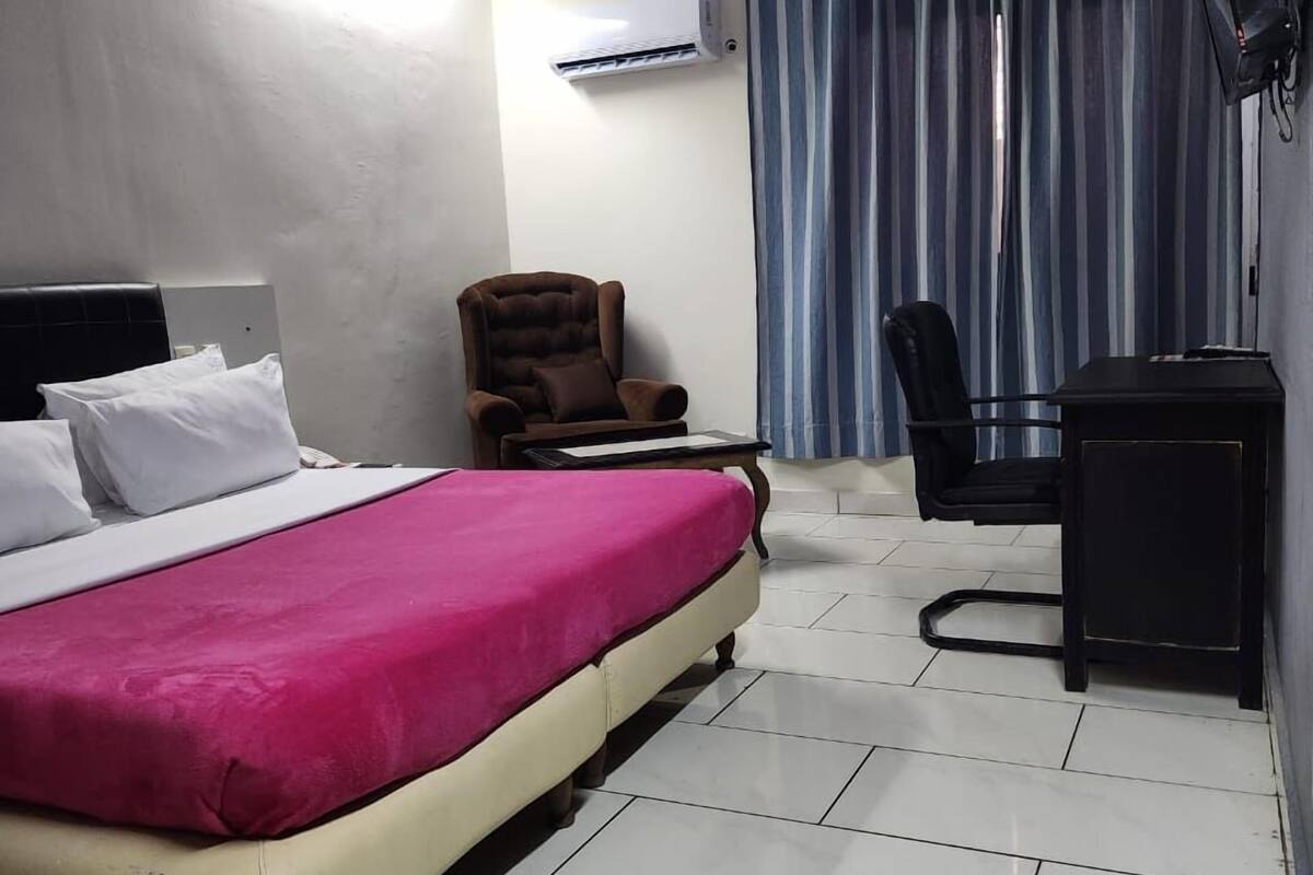 A hotel room with a bed, a desk, a chair, a TV, and an air conditioning unit.