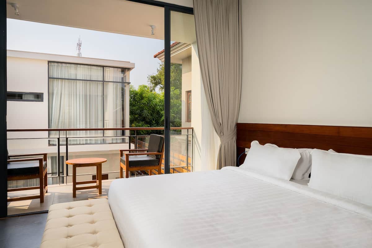 A hotel room with a large bed, a balcony with a table and chairs, and a view of greenery and buildings.