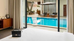 A hotel room with a large window overlooking a swimming pool, a wooden side table, and a black handbag on the bed.