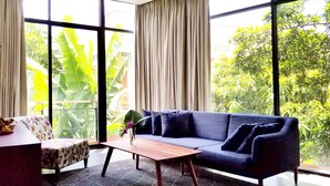 A modern living room with a blue sofa, a wooden coffee table, and a patterned rug. Large windows offer a view of lush greenery outside. - Angkor Siesta Villa (Siem Reap)