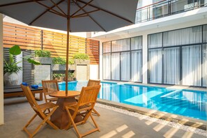 A modern outdoor pool area with a wooden table and chairs, surrounded by a wooden fence and large windows.