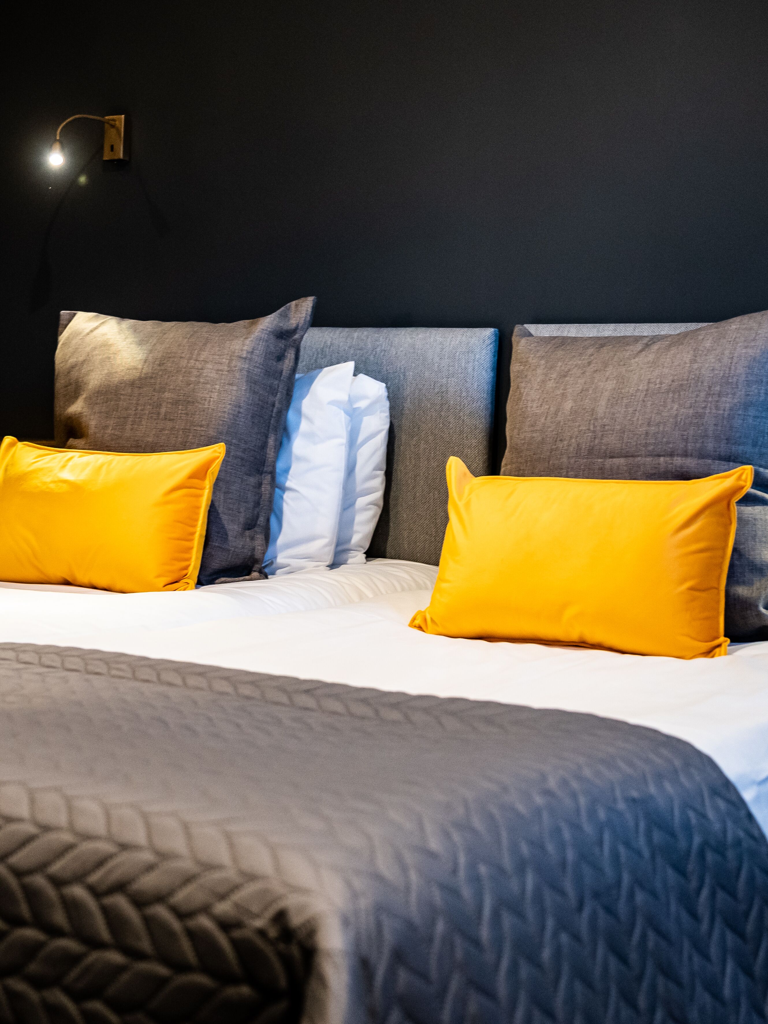 A neatly made bed with a grey quilt and two yellow pillows, a wall-mounted light, and a dark wall.