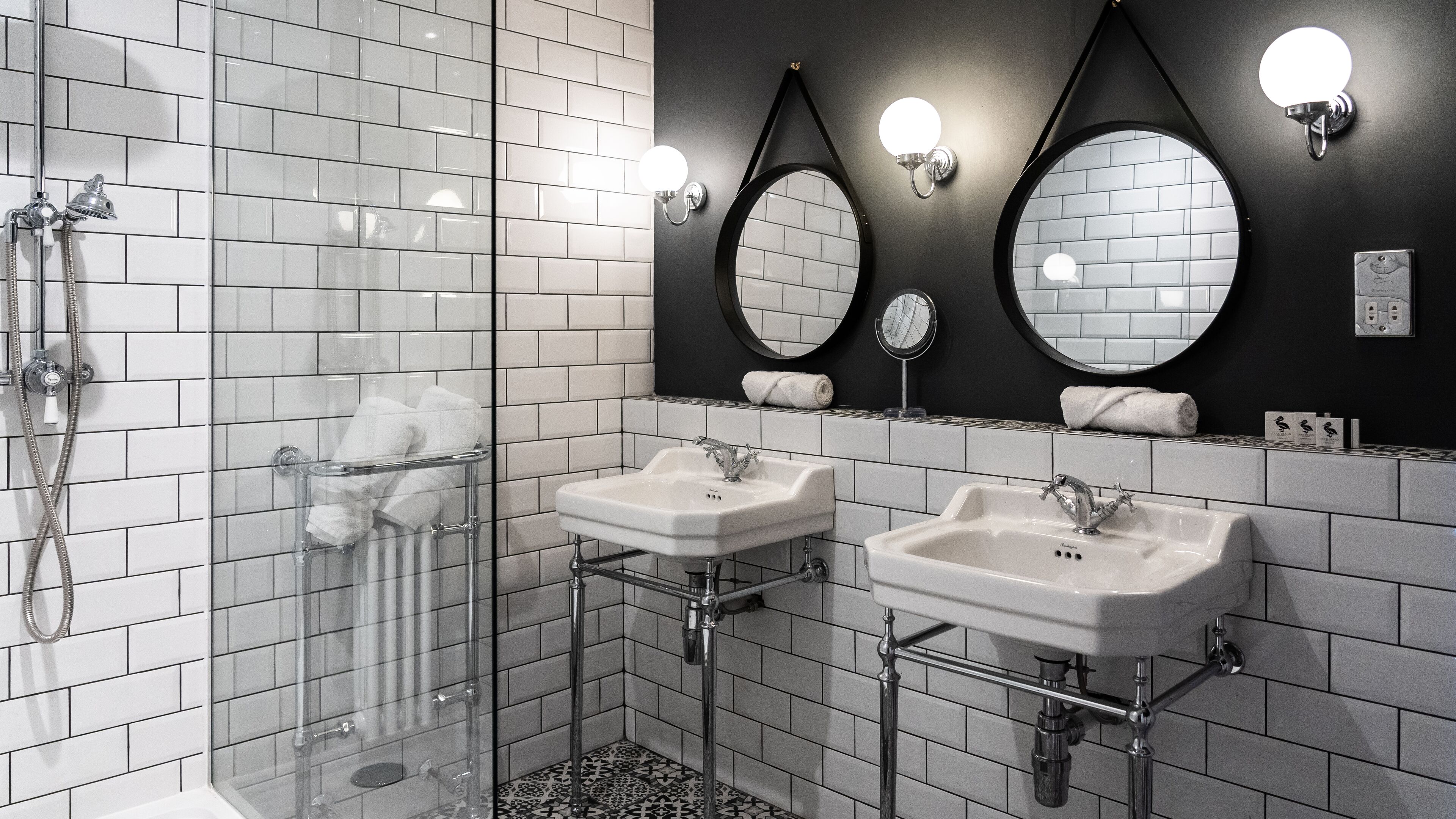 Une salle de bain moderne avec des murs recouverts de carreaux blancs, deux vasques, une douche en verre et des miroirs fixés au mur.