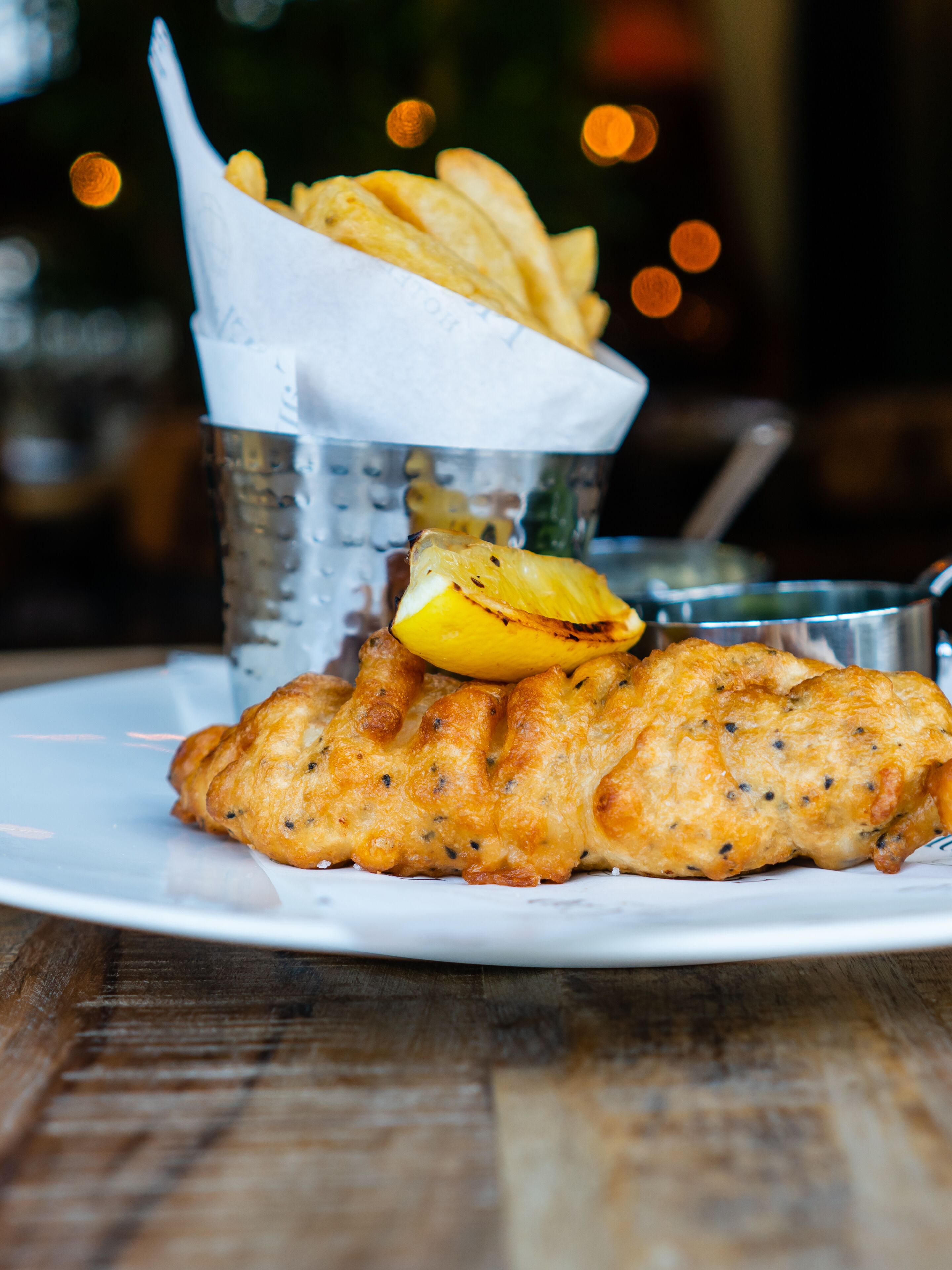 Un plato con un filete de pescado, papas fritas, un gajo de limón y una bebida en un vaso metálico.