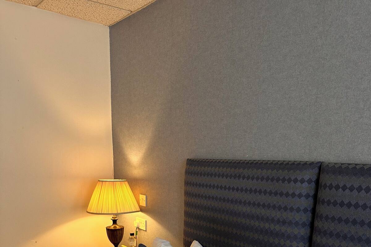 A neatly made bed with white linens and dark patterned pillows, a bedside lamp, and a small table with a cup and a bottle.