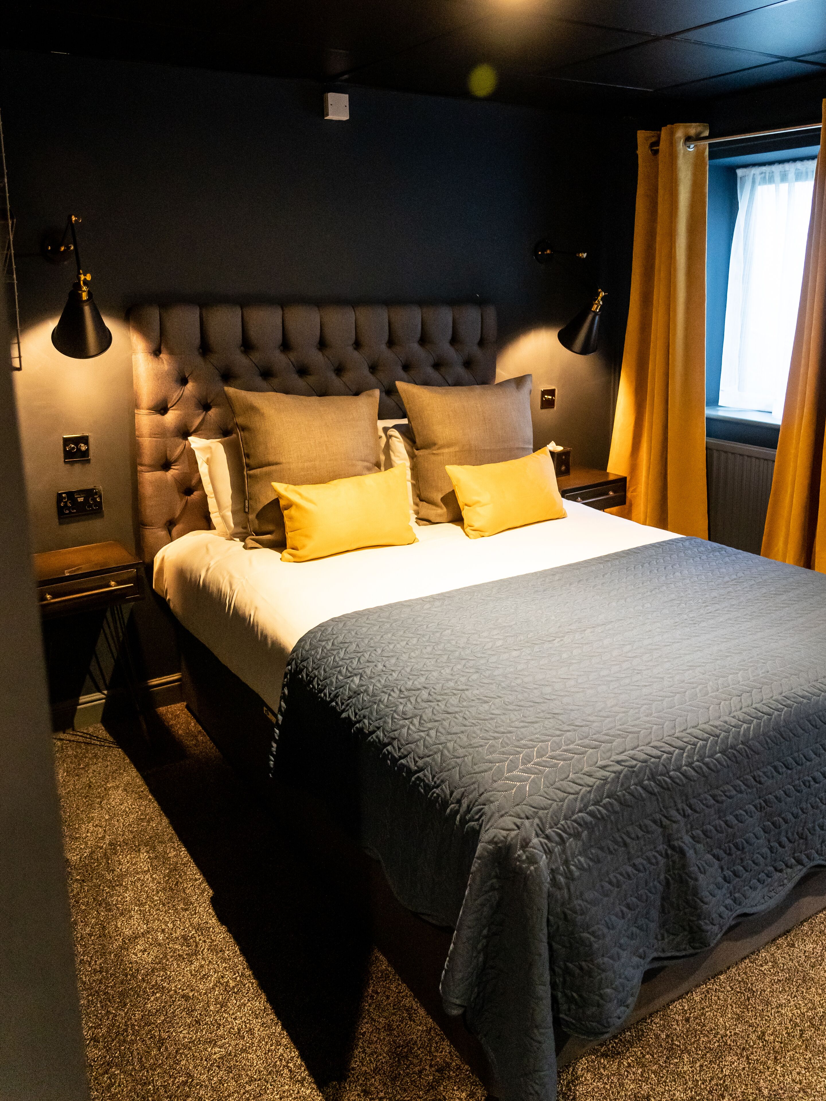 A bedroom with a tufted headboard, a bed with a quilted cover, two bedside lamps, and a window with curtains.