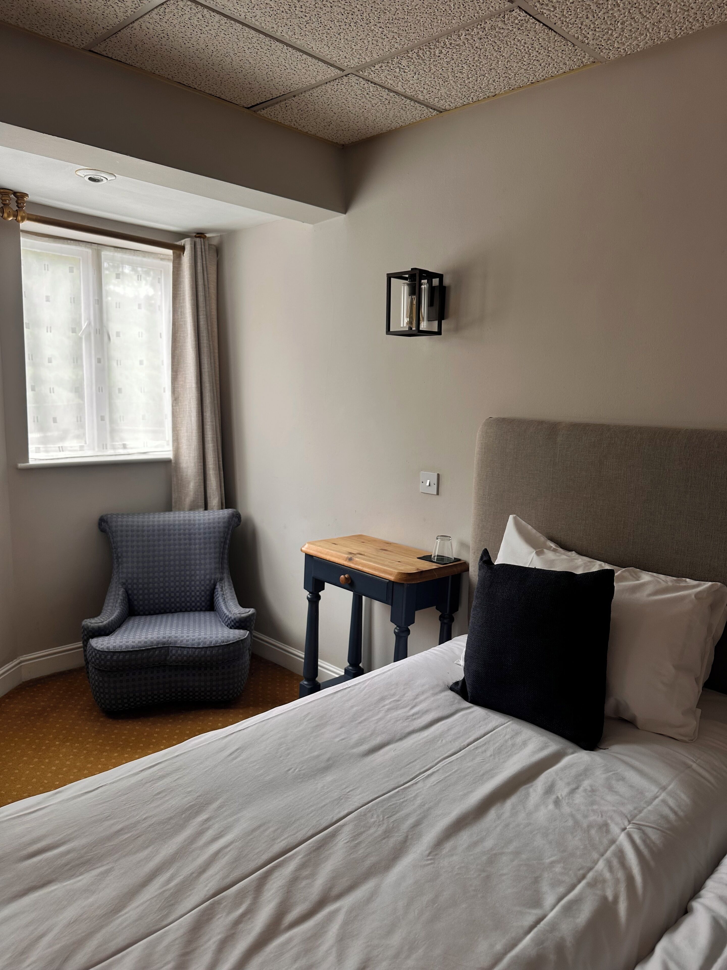 A hotel room with a bed, a chair, a small table, and a wall-mounted light fixture.