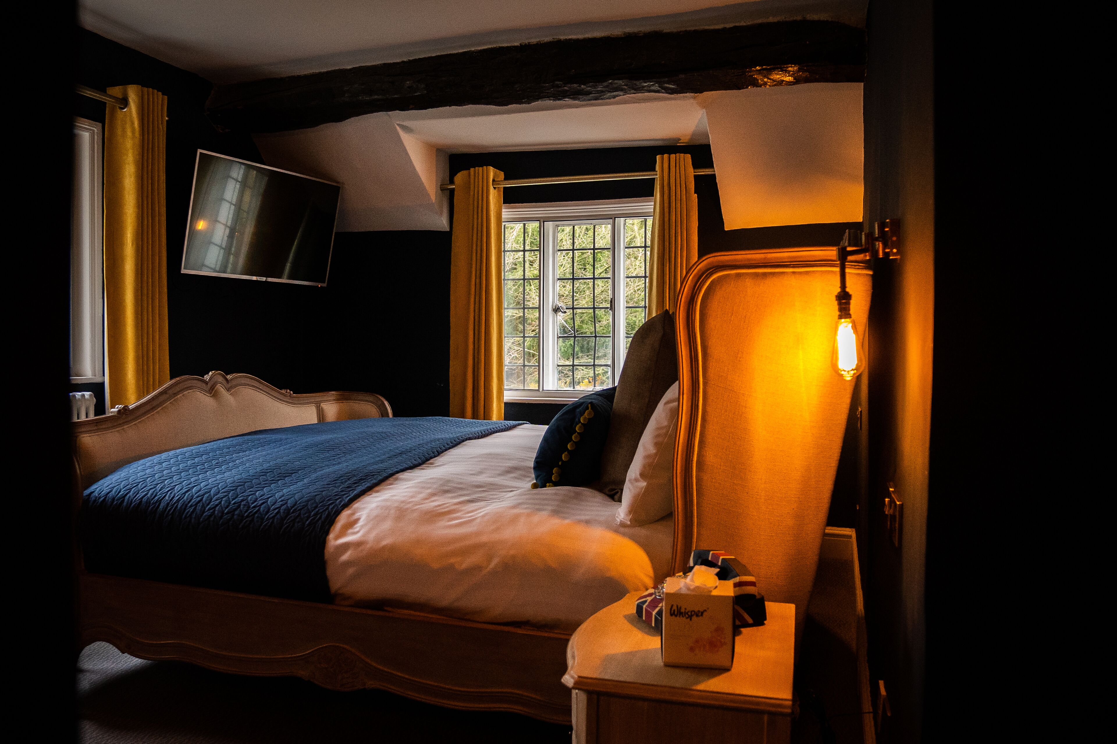 A hotel room with a bed, a bedside table with a lamp, a TV mounted on the wall, and a window with curtains.