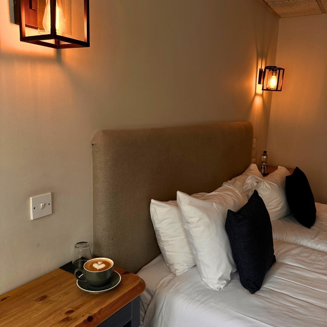 A hotel room with a bed, pillows, a bedside table with a cup of coffee, and wall-mounted lights.