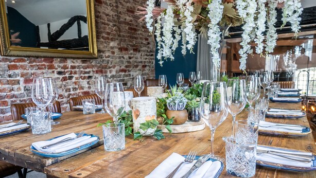 A long wooden table set for a meal with clear glassware, blue napkins, and a rustic brick wall in the background. - The Bridge (Macclesfield)