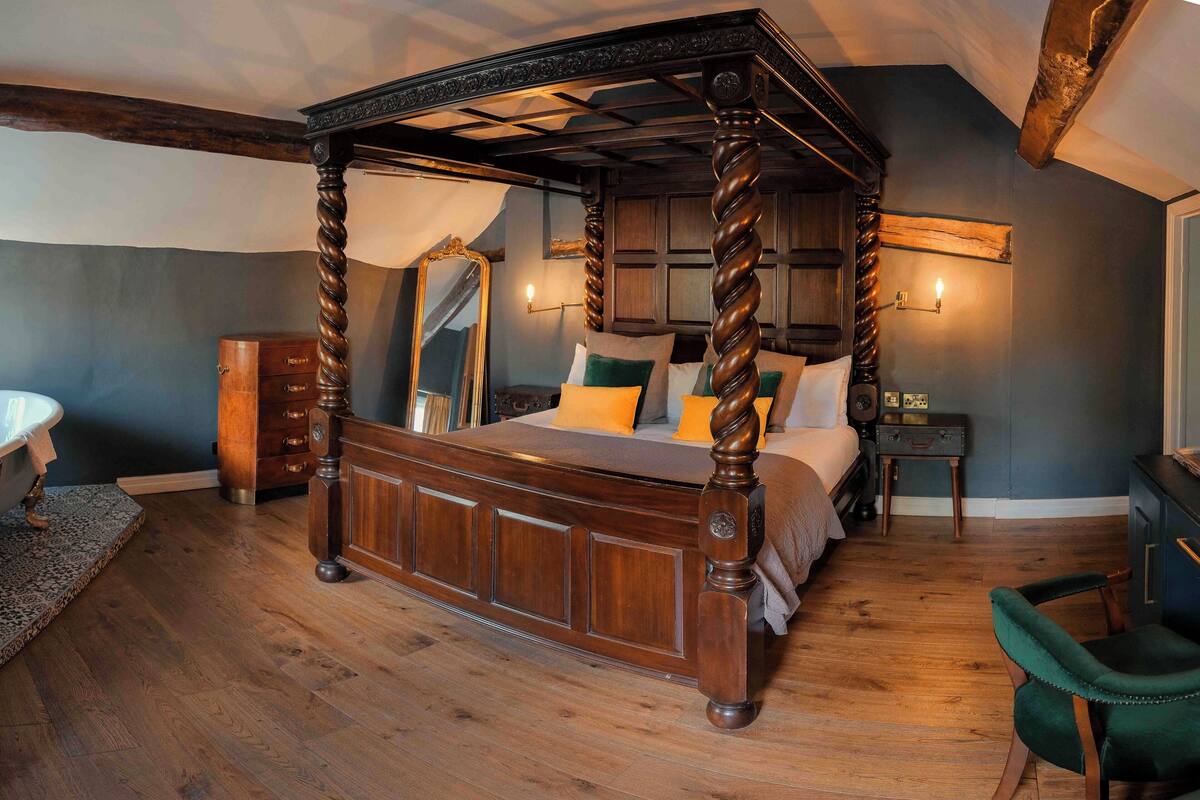 A four-poster bed with a dark wooden frame and carved details, a mirror on the wall, and a small bedside table with a lamp.