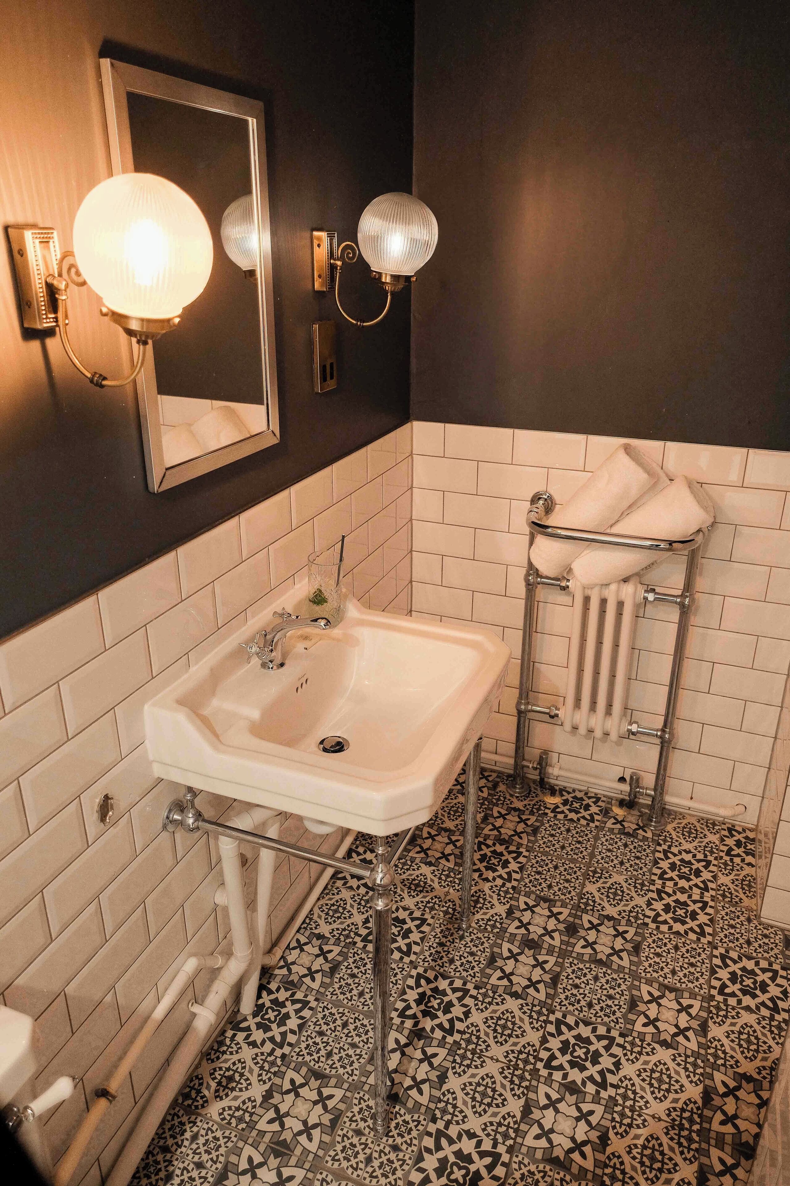 A bathroom with a white sink, a mirror, two wall-mounted lights, a towel rack, and patterned floor tiles.