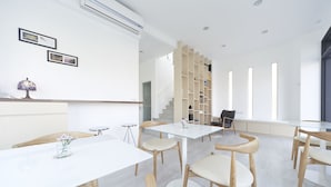 Une pièce moderne et bien éclairée, avec des murs blancs, une table en verre et des chaises en bois. Des tableaux encadrés ornent le mur, une lampe se trouve sur une table d’appoint, et un escalier mène à un autre niveau.
