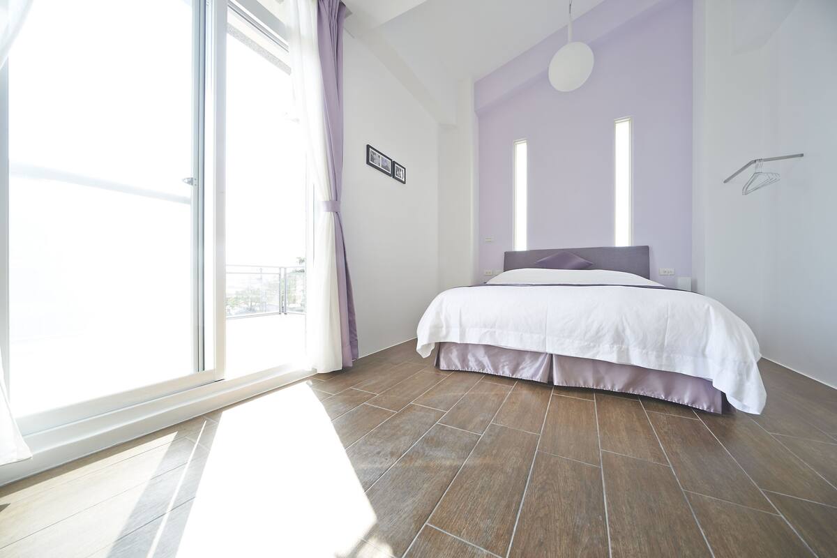 A bedroom with a large window, a bed with a white and purple bedspread, and a brown tiled floor.