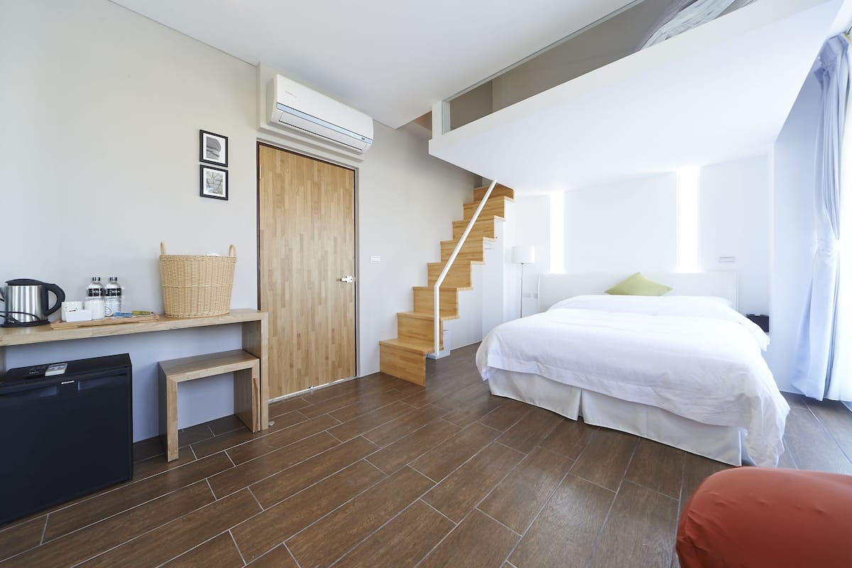 A modern hotel room with a wooden desk, a bed, a staircase, and a large window.