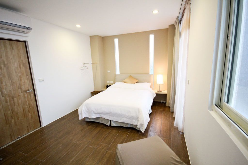 Una habitación de hotel con una cama, mesitas de noche, una lámpara colgante, una ventana con cortinas y una puerta de madera.