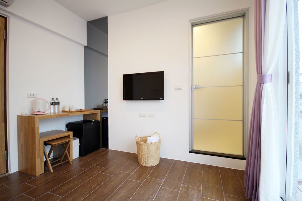 Ein gemütlicher Raum mit Schreibtisch aus Holz, einem an der Wand befestigten schwarzen Fernseher, einer kleinen Mülltonne und einem Korbwäscher.