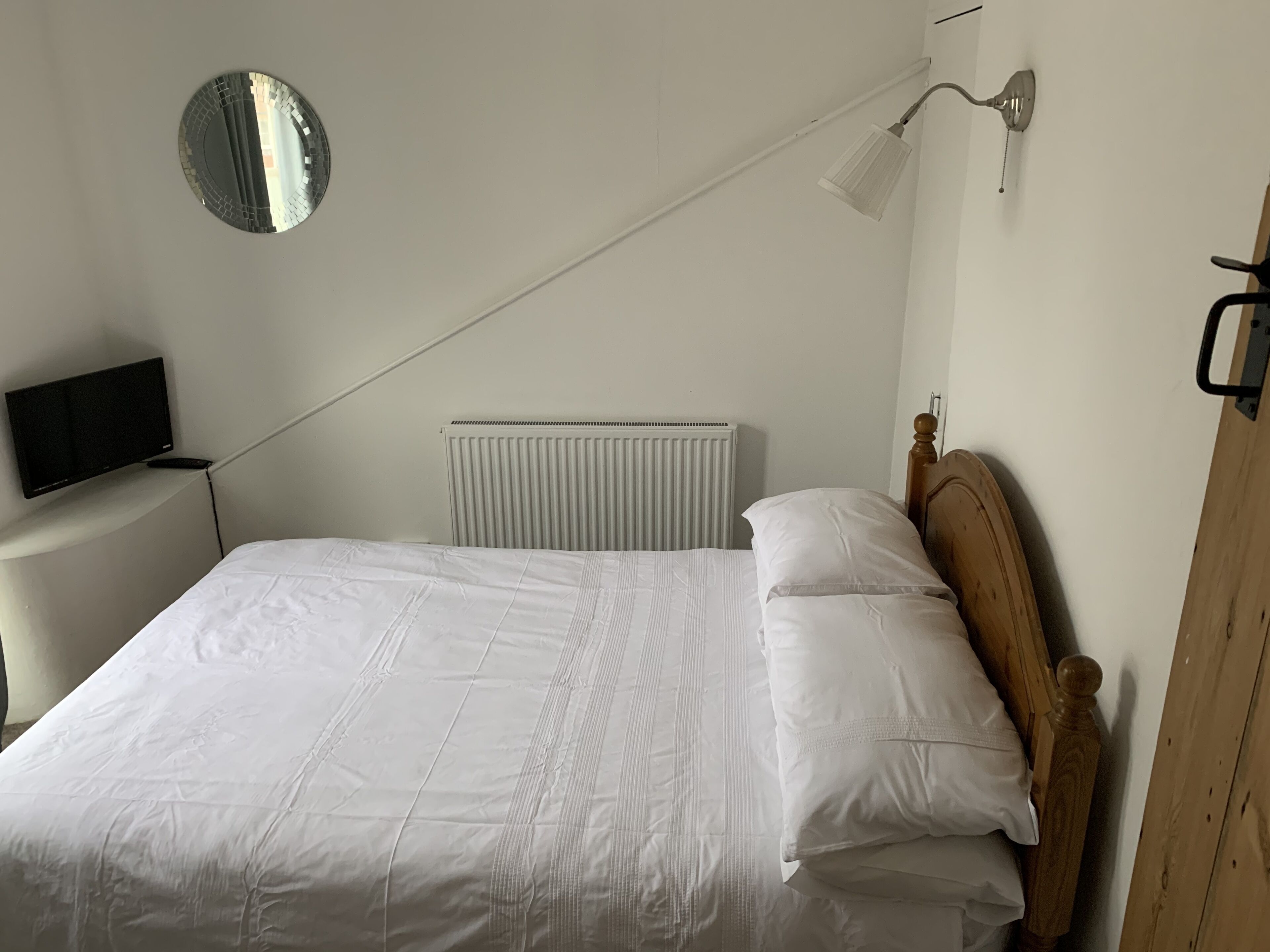 Un letto rifatto con lenzuola bianche, uno specchio rotondo al muro, un comodino con una TV a schermo piatto e un termosifone sopra il letto.