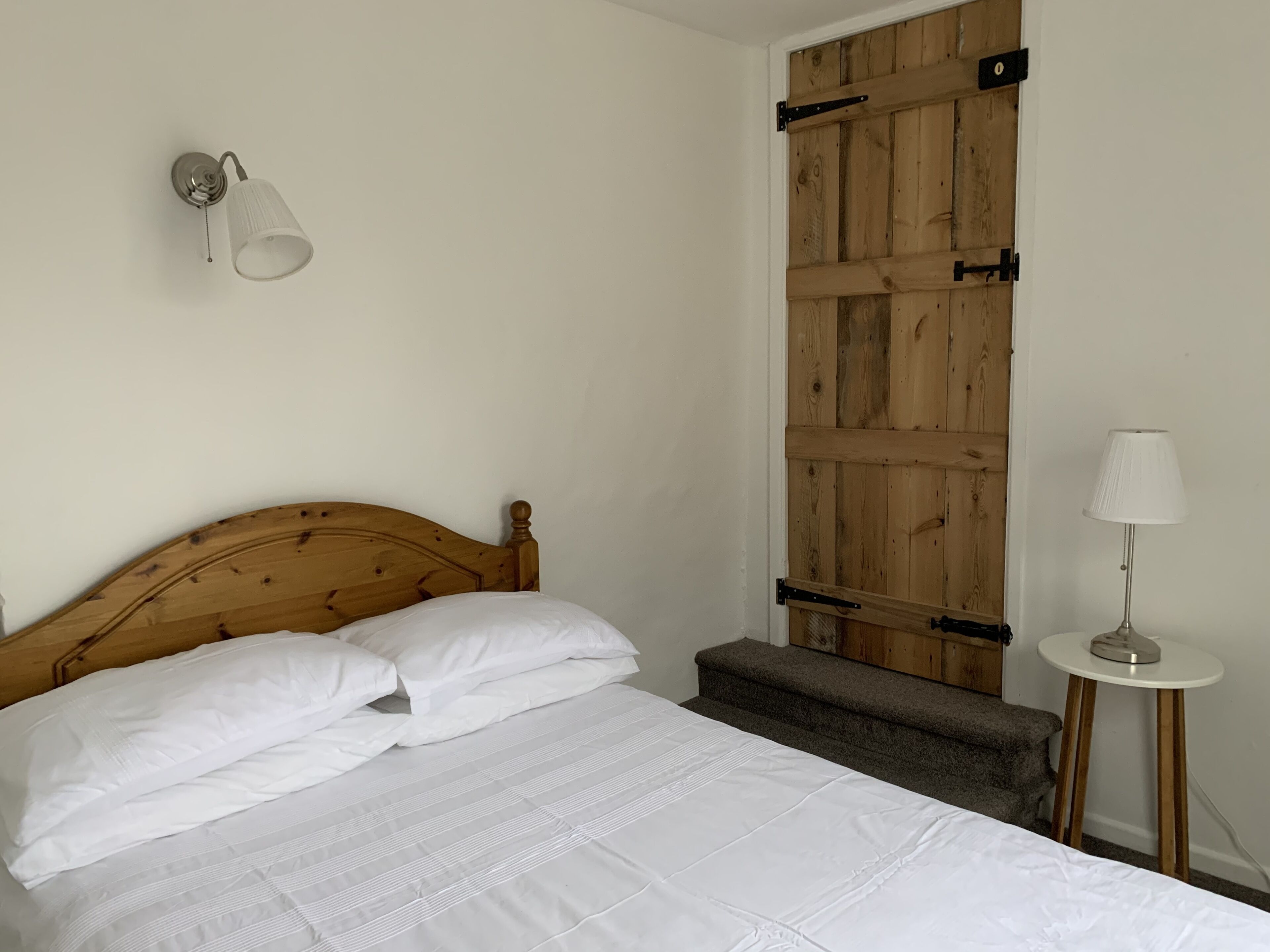 A bedroom with a wooden headboard, a bed with white bedding, a small bedside table with a lamp, and a wooden door with metal hinges.