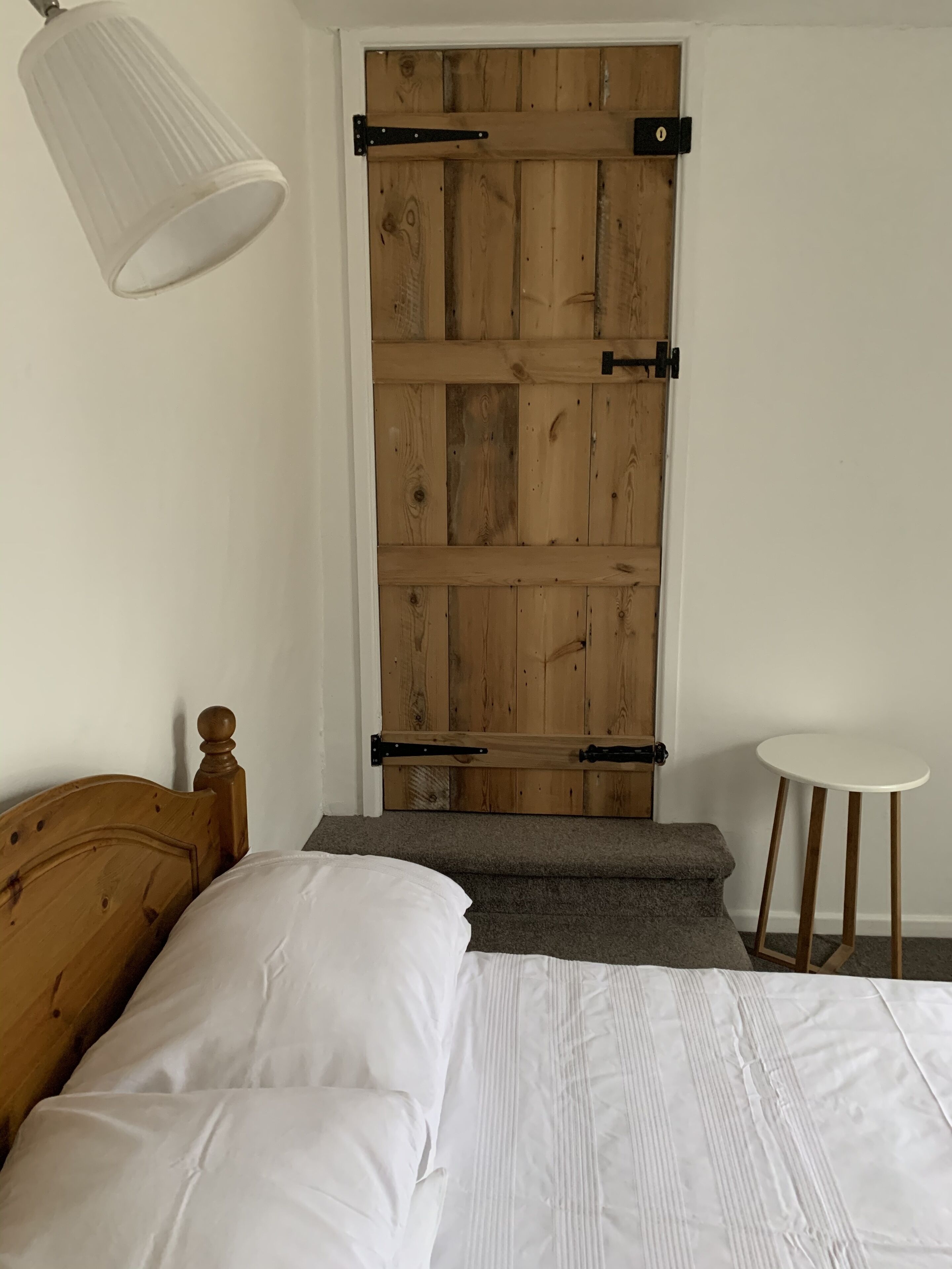 A bedroom with a wooden door, a bed with white bedding, a small white table, and a hanging light fixture.