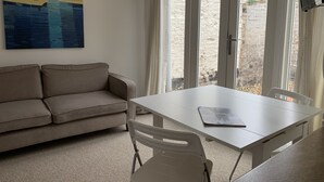A room with a beige sofa, a white table, and a large window with curtains. - Bell Cottage (Bury St Edmunds)