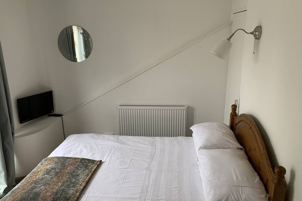 Een net opgemaakt bed met witte lakens, een houten hoofdbord, een bedlampje, een aan de muur bevestigde televisie en een radiator.