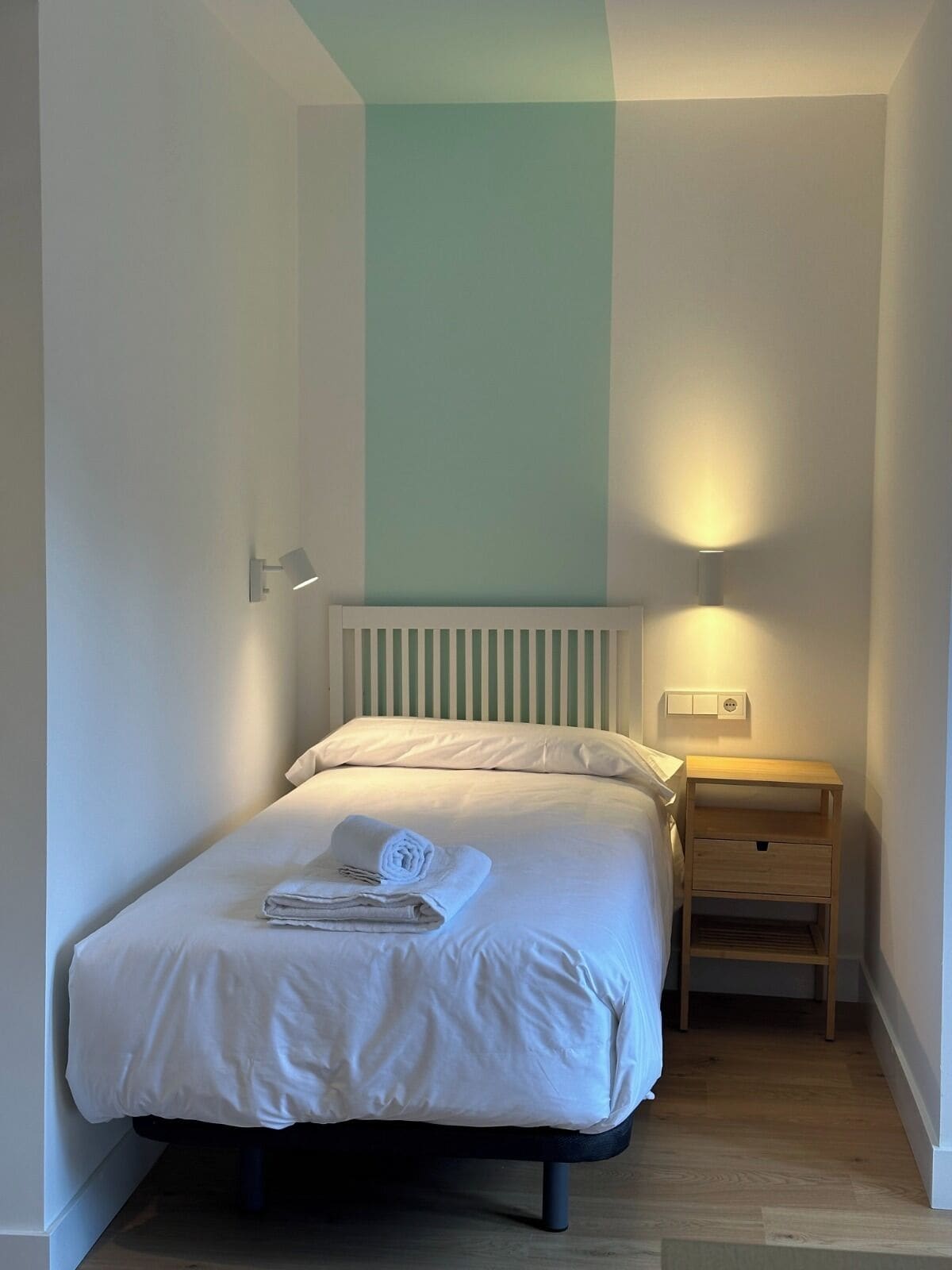Una cama pequeña y ordenada con ropa de cama blanca y una toalla doblada, una mesita de noche con lámpara, y un aplique de pared.