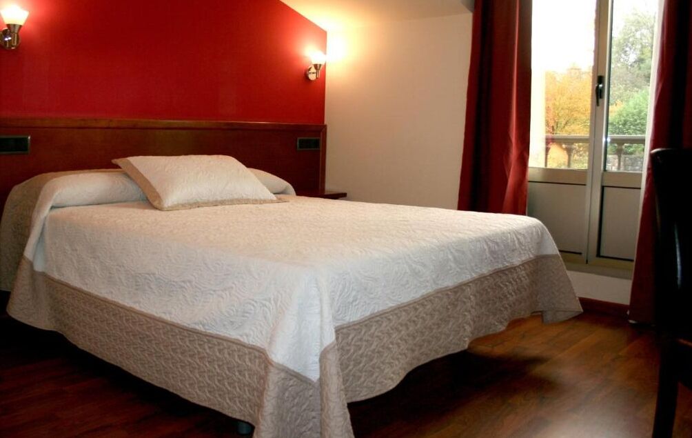 A bed with a white bedspread and two pillows, a wooden headboard, a nightstand, a window with red curtains, and a view of trees outside.